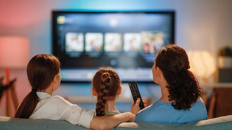 Mãe e duas filhas a ver televisão sentadas no sofá; vista de trás